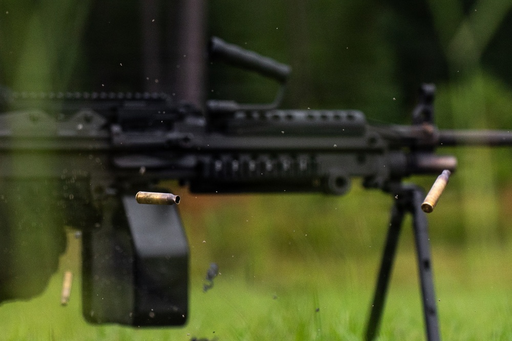 A high-speed photograph captures a light machine gun mounted on a bipod in the process of firing, with two brass cartridge casings visibly ejected mid-air. The weapon appears to be an FN M249 SAW or similar squad automatic weapon, identifiable by its box magazine, carry handle, and Picatinny rail system. Water droplets or debris particles are also visible suspended in the air around the weapon.