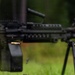 A high-speed photograph captures a light machine gun mounted on a bipod in the process of firing, with two brass cartridge casings visibly ejected mid-air. The weapon appears to be an FN M249 SAW or similar squad automatic weapon, identifiable by its box magazine, carry handle, and Picatinny rail system. Water droplets or debris particles are also visible suspended in the air around the weapon.
