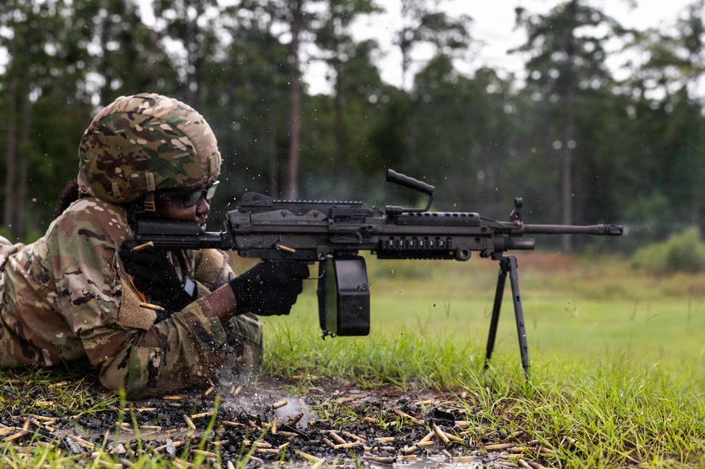 A soldier in MultiCam/OCP camouflage uniform is firing an M249 Squad Automatic Weapon (SAW) from a prone position on wet ground. The weapon is mounted on its bipod and appears to be actively firing, as evidenced by spent brass casings scattered around the shooter. The setting appears to be a military training range in a wooded area during or after rainfall.