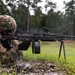 A soldier in MultiCam/OCP camouflage uniform is firing an M249 Squad Automatic Weapon (SAW) from a prone position on wet ground. The weapon is mounted on its bipod and appears to be actively firing, as evidenced by spent brass casings scattered around the shooter. The setting appears to be a military training range in a wooded area during or after rainfall.