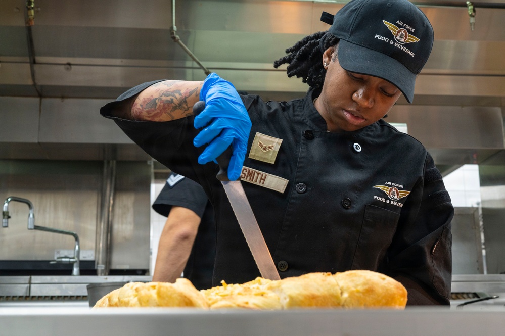 A U.S. Air Force Food &amp; Beverage specialist, identified by the name tape 'SMITH', is working in a military kitchen preparing baked goods. The individual is wearing a black chef's uniform with rank insignia, blue nitrile gloves, and an Air Force Food &amp; Beverage cap. They are using tongs or a knife to work with what appears to be freshly baked cheese-topped bread rolls.
