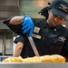 A U.S. Air Force Food &amp; Beverage specialist, identified by the name tape 'SMITH', is working in a military kitchen preparing baked goods. The individual is wearing a black chef's uniform with rank insignia, blue nitrile gloves, and an Air Force Food &amp; Beverage cap. They are using tongs or a knife to work with what appears to be freshly baked cheese-topped bread rolls.