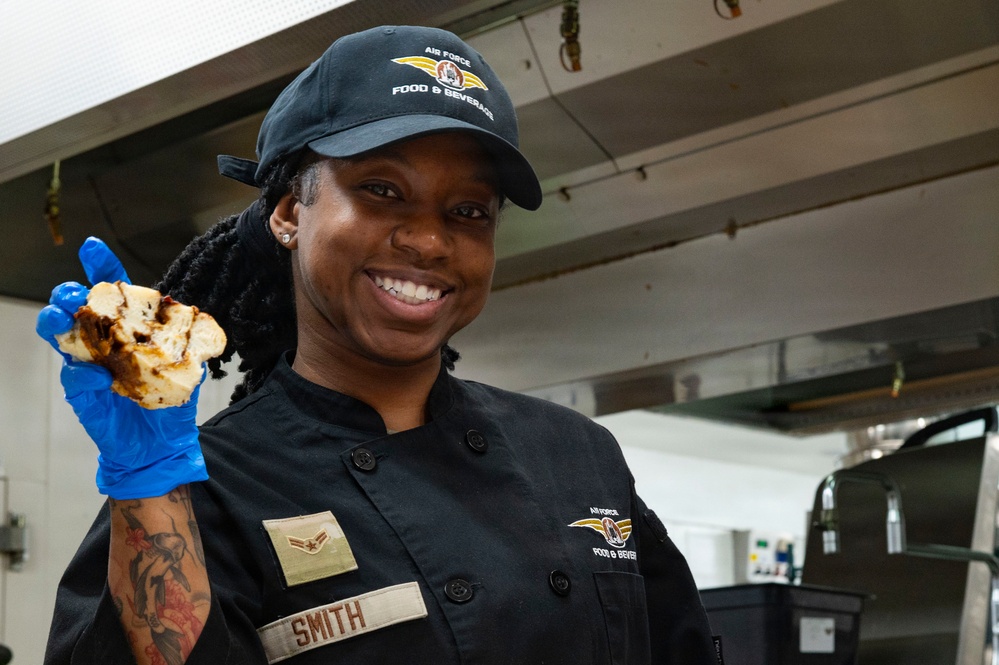 A U.S. Air Force Food &amp; Beverage specialist named Smith is photographed in a military kitchen setting. She is smiling and holding up a piece of cooked food while wearing blue latex gloves and a black chef's uniform. Her name tag, rank insignia (Airman First Class), and Air Force Food &amp; Beverage patches are clearly visible.