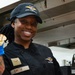 A U.S. Air Force Food &amp; Beverage specialist named Smith is photographed in a military kitchen setting. She is smiling and holding up a piece of cooked food while wearing blue latex gloves and a black chef's uniform. Her name tag, rank insignia (Airman First Class), and Air Force Food &amp; Beverage patches are clearly visible.
