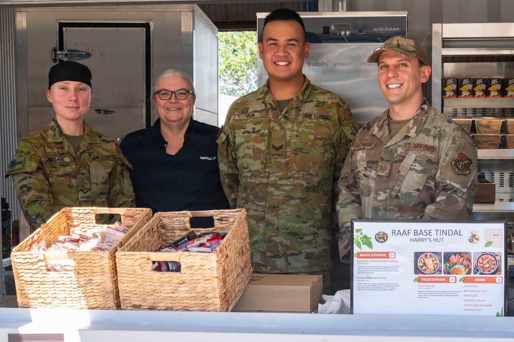 Four individuals pose together at a food service counter at RAAF Base Tindal's 'Harry's Hut' canteen. The group includes three military personnel in camouflage uniforms (two Australian Air Force and one U.S. Air Force) and one civilian woman wearing a 'Ventia' branded black polo shirt. A menu board, wicker baskets with snacks, and commercial kitchen equipment are visible in the background.