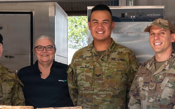 Four individuals pose together at a food service counter at RAAF Base Tindal's 'Harry's Hut' canteen. The group includes three military personnel in camouflage uniforms (two Australian Air Force and one U.S. Air Force) and one civilian woman wearing a 'Ventia' branded black polo shirt. A menu board, wicker baskets with snacks, and commercial kitchen equipment are visible in the background.
