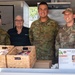 Four individuals pose together at a food service counter at RAAF Base Tindal's 'Harry's Hut' canteen. The group includes three military personnel in camouflage uniforms (two Australian Air Force and one U.S. Air Force) and one civilian woman wearing a 'Ventia' branded black polo shirt. A menu board, wicker baskets with snacks, and commercial kitchen equipment are visible in the background.