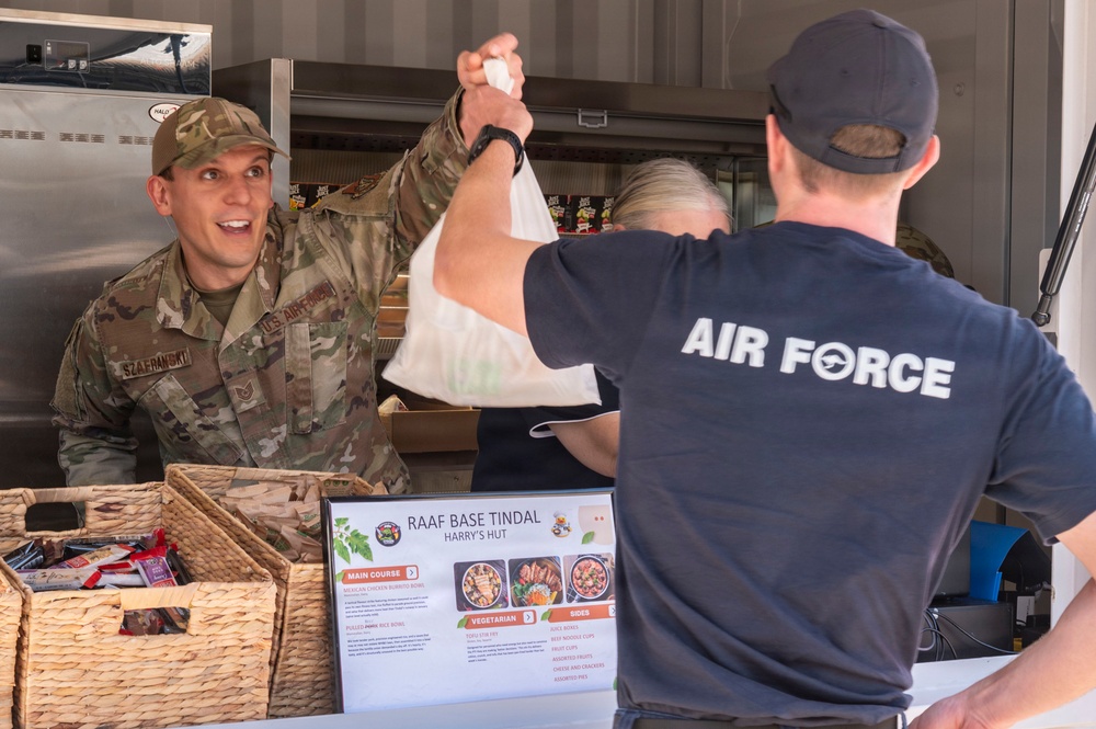 A U.S. Air Force service member in camouflage uniform is receiving a food bag at RAAF Base Tindal's 'Harry's Hut' food service station. Another individual wearing an 'AIR FORCE' t-shirt is handing over the bag across a counter. The scene depicts a military dining or food distribution operation at an Australian Royal Air Force base.
