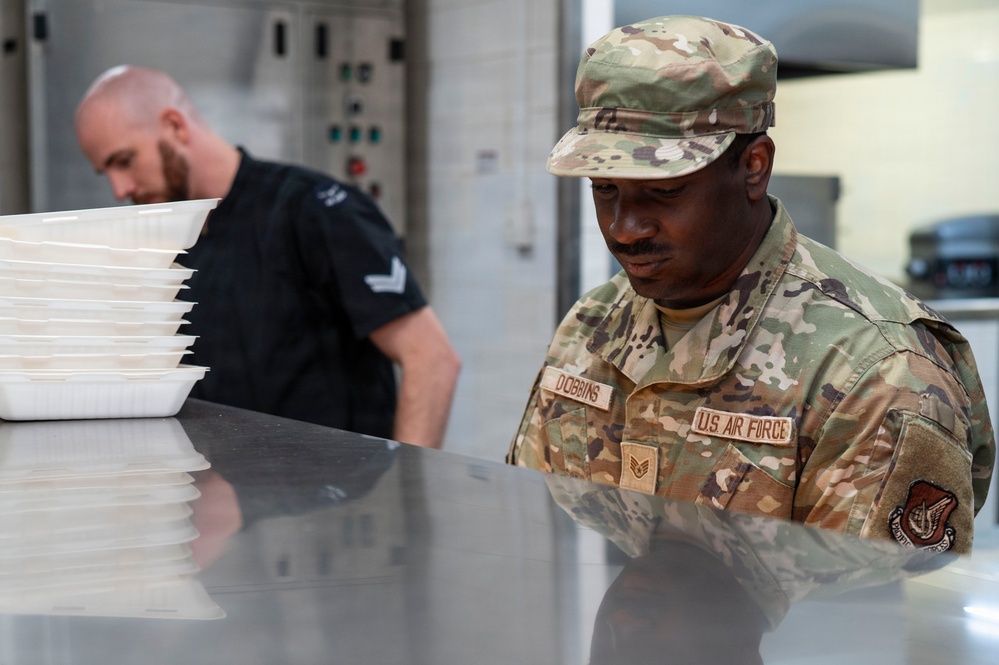 Two individuals are working in what appears to be a military kitchen or food service facility. The foreground figure is a U.S. Air Force Staff Sergeant named Dobbins in OCP (Operational Camouflage Pattern) uniform, focused on a stainless steel work surface. A second individual in a black uniform with rank insignia is handling stacked white food trays in the background.