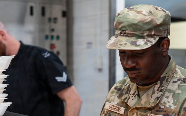 Two individuals are working in what appears to be a military kitchen or food service facility. The foreground figure is a U.S. Air Force Staff Sergeant named Dobbins in OCP (Operational Camouflage Pattern) uniform, focused on a stainless steel work surface. A second individual in a black uniform with rank insignia is handling stacked white food trays in the background.