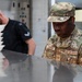 Two individuals are working in what appears to be a military kitchen or food service facility. The foreground figure is a U.S. Air Force Staff Sergeant named Dobbins in OCP (Operational Camouflage Pattern) uniform, focused on a stainless steel work surface. A second individual in a black uniform with rank insignia is handling stacked white food trays in the background.
