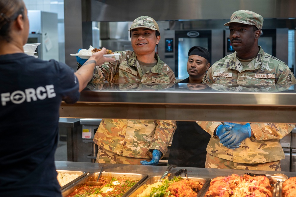 Two U.S. Air Force personnel in OCP (Operational Camouflage Pattern) uniforms are serving food at a military dining facility (DFAC). The airmen, identified by name tapes as 'RIMAL' and 'DOBBINS,' are wearing blue food-service gloves and working behind a cafeteria-style serving counter. A civilian or staff member in a dark 'FORCE' t-shirt is receiving a plate of food from Airman Rimal.