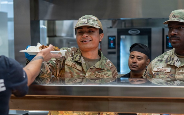 Two U.S. Air Force personnel in OCP (Operational Camouflage Pattern) uniforms are serving food at a military dining facility (DFAC). The airmen, identified by name tapes as 'RIMAL' and 'DOBBINS,' are wearing blue food-service gloves and working behind a cafeteria-style serving counter. A civilian or staff member in a dark 'FORCE' t-shirt is receiving a plate of food from Airman Rimal.
