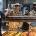 Two U.S. Air Force personnel in OCP (Operational Camouflage Pattern) uniforms are serving food at a military dining facility (DFAC). The airmen, identified by name tapes as 'RIMAL' and 'DOBBINS,' are wearing blue food-service gloves and working behind a cafeteria-style serving counter. A civilian or staff member in a dark 'FORCE' t-shirt is receiving a plate of food from Airman Rimal.