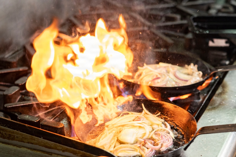 A dramatic flambé cooking technique is being performed on a commercial gas stove, with large orange and yellow flames rising from a cast iron skillet. Two cast iron pans are visible on the burner grates, both containing what appears to be sautéed onions. The intense flames suggest alcohol has been added to the pan intentionally or oil has ignited during high-heat cooking.
