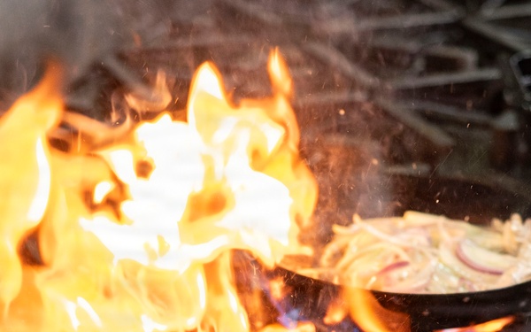 A dramatic flambé cooking technique is being performed on a commercial gas stove, with large orange and yellow flames rising from a cast iron skillet. Two cast iron pans are visible on the burner grates, both containing what appears to be sautéed onions. The intense flames suggest alcohol has been added to the pan intentionally or oil has ignited during high-heat cooking.