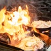 A dramatic flambé cooking technique is being performed on a commercial gas stove, with large orange and yellow flames rising from a cast iron skillet. Two cast iron pans are visible on the burner grates, both containing what appears to be sautéed onions. The intense flames suggest alcohol has been added to the pan intentionally or oil has ignited during high-heat cooking.