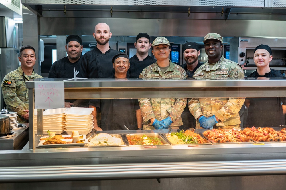 A group of approximately nine individuals pose together behind a military dining facility (DFAC) serving line. The group includes U.S. Air Force personnel in MultiCam uniforms, a U.S. Army soldier, and civilian kitchen staff dressed in black chef attire. A sign reading 'MEAL TICKETS' is visible at the serving counter, which displays multiple food trays.