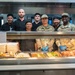 A group of approximately nine individuals pose together behind a military dining facility (DFAC) serving line. The group includes U.S. Air Force personnel in MultiCam uniforms, a U.S. Army soldier, and civilian kitchen staff dressed in black chef attire. A sign reading 'MEAL TICKETS' is visible at the serving counter, which displays multiple food trays.