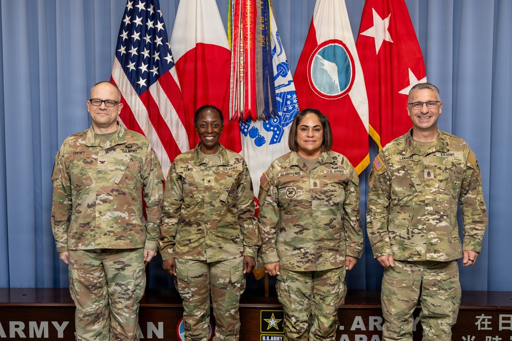 Four U.S. Army personnel in OCP (Operational Camouflage Pattern) uniforms pose for a formal group photograph in front of multiple flags including the U.S. flag, Japanese flag, and U.S. Army command flags. Name tapes visible on uniforms read Cheney, Lewis, Quintero, and Lancet from left to right. The setting appears to be an official U.S. Army Japan headquarters or command facility.