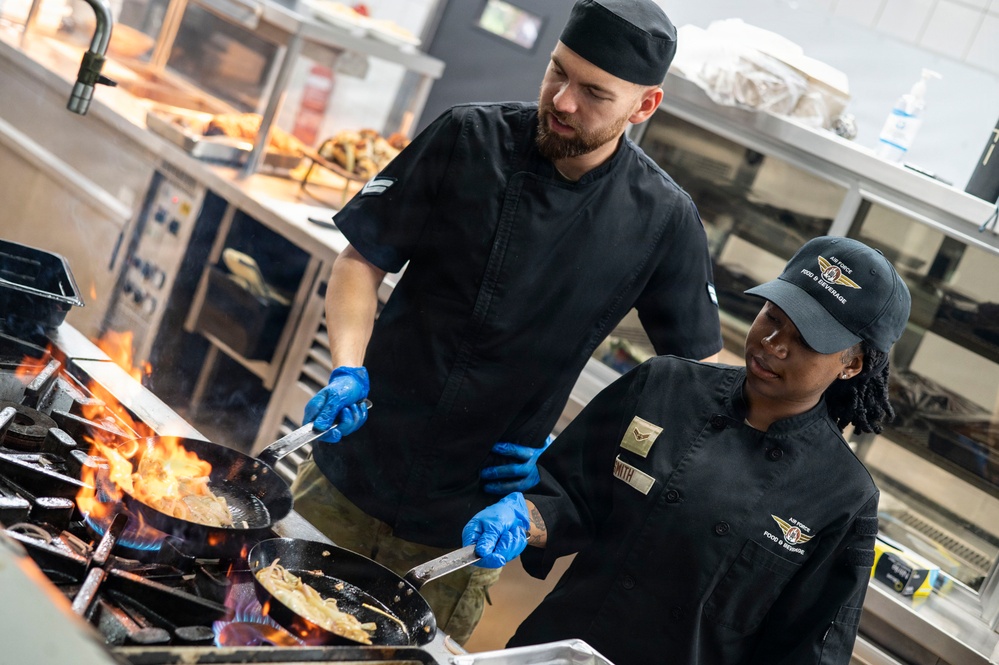 Two kitchen workers dressed in black chef uniforms and blue latex gloves are cooking over open-flame gas burners in a professional kitchen. The person on the right is wearing a cap and uniform patch reading 'Air Force Food &amp; Beverage,' suggesting this is a military dining facility. Both individuals appear to be actively sautéing food in pans with visible flames.