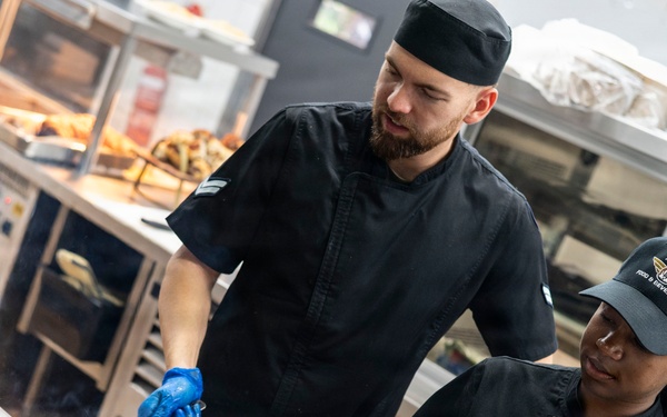 Two kitchen workers dressed in black chef uniforms and blue latex gloves are cooking over open-flame gas burners in a professional kitchen. The person on the right is wearing a cap and uniform patch reading 'Air Force Food &amp; Beverage,' suggesting this is a military dining facility. Both individuals appear to be actively sautéing food in pans with visible flames.