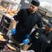 Two kitchen workers dressed in black chef uniforms and blue latex gloves are cooking over open-flame gas burners in a professional kitchen. The person on the right is wearing a cap and uniform patch reading 'Air Force Food &amp; Beverage,' suggesting this is a military dining facility. Both individuals appear to be actively sautéing food in pans with visible flames.