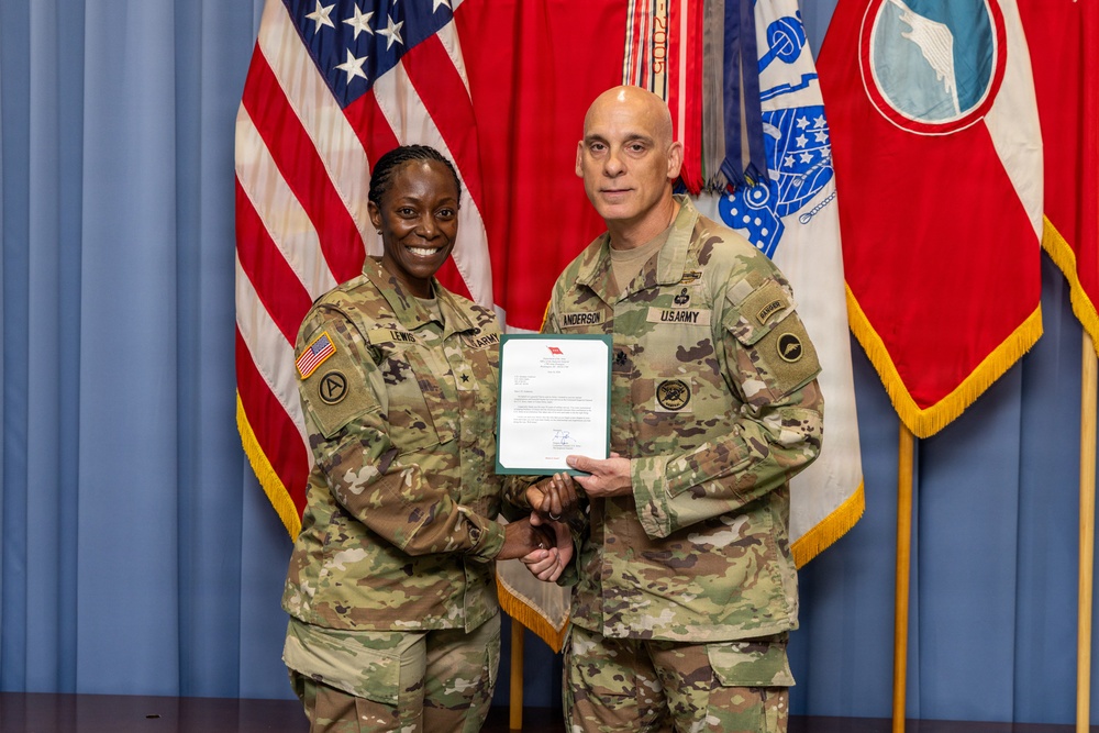 Two U.S. Army soldiers in OCP (Operational Camouflage Pattern) uniforms pose together holding an official document during what appears to be a military ceremony. The name tapes visible read 'LEWIS' on the left and 'ANDERSON' on the right. They are standing in front of U.S. and military unit flags against a blue backdrop.