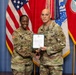 Two U.S. Army soldiers in OCP (Operational Camouflage Pattern) uniforms pose together holding an official document during what appears to be a military ceremony. The name tapes visible read 'LEWIS' on the left and 'ANDERSON' on the right. They are standing in front of U.S. and military unit flags against a blue backdrop.