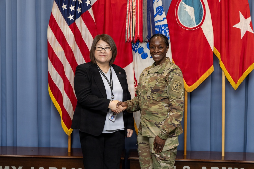 Two individuals are shaking hands in a formal military setting. One person is wearing a U.S. Army camouflage uniform (OCP pattern) and the other is dressed in civilian business attire with a lanyard and badge. Multiple flags, including the U.S. flag and military unit flags, are displayed in the background.