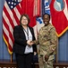 Two individuals are shaking hands in a formal military setting. One person is wearing a U.S. Army camouflage uniform (OCP pattern) and the other is dressed in civilian business attire with a lanyard and badge. Multiple flags, including the U.S. flag and military unit flags, are displayed in the background.
