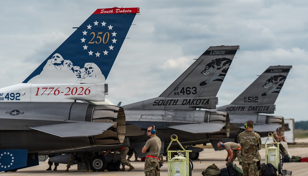 Three F-16 Fighting Falcon jets from the South Dakota Air National Guard are parked on a flight line tarmac. The foreground aircraft features a special commemorative paint scheme celebrating the United States 250th anniversary (1776-2026) with Mount Rushmore imagery and '250' surrounded by stars on the tail. Ground crew personnel in military uniforms are performing maintenance and servicing operations on the aircraft.