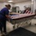 Three workers in blue scrubs or uniforms are carefully draping an American flag over a casket on a wheeled mortuary gurney inside what appears to be a mortuary or medical examiner facility. The scene depicts a dignified military or veteran honors preparation process. A second flag-draped casket is visible in the background on a table.