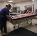 Three workers in blue scrubs or uniforms are carefully draping an American flag over a casket on a wheeled mortuary gurney inside what appears to be a mortuary or medical examiner facility. The scene depicts a dignified military or veteran honors preparation process. A second flag-draped casket is visible in the background on a table.