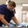A person wearing blue scrubs, safety goggles, an N95 respirator mask, and blue nitrile gloves is working at a table in what appears to be a laboratory or clinical facility. They are handling an organic or biological specimen wrapped in blue disposable draping material. A second similarly dressed individual is visible in the background, suggesting a team-based procedural environment.