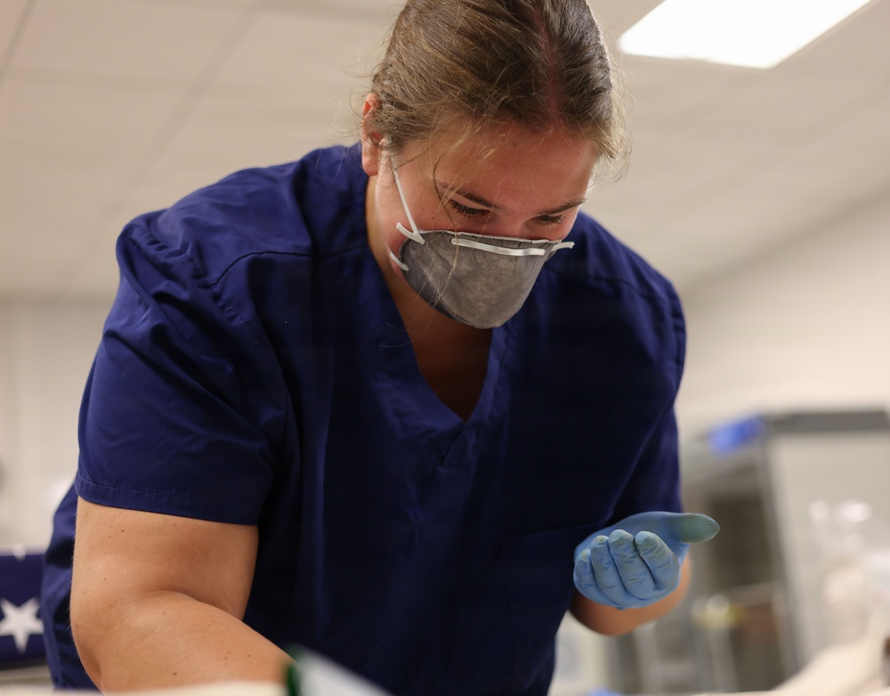 A healthcare or laboratory worker wearing blue scrubs, an N95 respirator mask, and blue nitrile gloves is leaning forward and focused on a task below the frame. The setting appears to be a clinical or laboratory environment with fluorescent overhead lighting. The worker appears engaged in a hands-on procedure or examination.