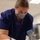 A healthcare or laboratory worker wearing blue scrubs, an N95 respirator mask, and blue nitrile gloves is leaning forward and focused on a task below the frame. The setting appears to be a clinical or laboratory environment with fluorescent overhead lighting. The worker appears engaged in a hands-on procedure or examination.