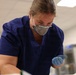 A healthcare or laboratory worker wearing blue scrubs, an N95 respirator mask, and blue nitrile gloves is leaning forward and focused on a task below the frame. The setting appears to be a clinical or laboratory environment with fluorescent overhead lighting. The worker appears engaged in a hands-on procedure or examination.
