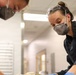 Two individuals wearing respiratory masks and protective gear are leaning over a work surface, focused on a task below the frame. The person on the right is also wearing safety goggles pushed up on their forehead and blue nitrile gloves. The setting appears to be an indoor facility with a corridor visible in the background.