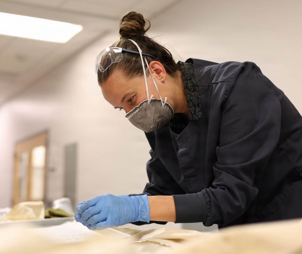 A professional, likely a conservator or archivist, is carefully examining fragile documents or textile materials on a work table. She is wearing appropriate personal protective equipment including blue nitrile gloves, an N95-style respirator mask, and safety goggles pushed up on her forehead. The setting appears to be a conservation lab or archival workspace with controlled lighting.