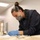 A professional, likely a conservator or archivist, is carefully examining fragile documents or textile materials on a work table. She is wearing appropriate personal protective equipment including blue nitrile gloves, an N95-style respirator mask, and safety goggles pushed up on her forehead. The setting appears to be a conservation lab or archival workspace with controlled lighting.