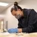 A professional, likely a conservator or archivist, is carefully examining fragile documents or textile materials on a work table. She is wearing appropriate personal protective equipment including blue nitrile gloves, an N95-style respirator mask, and safety goggles pushed up on her forehead. The setting appears to be a conservation lab or archival workspace with controlled lighting.