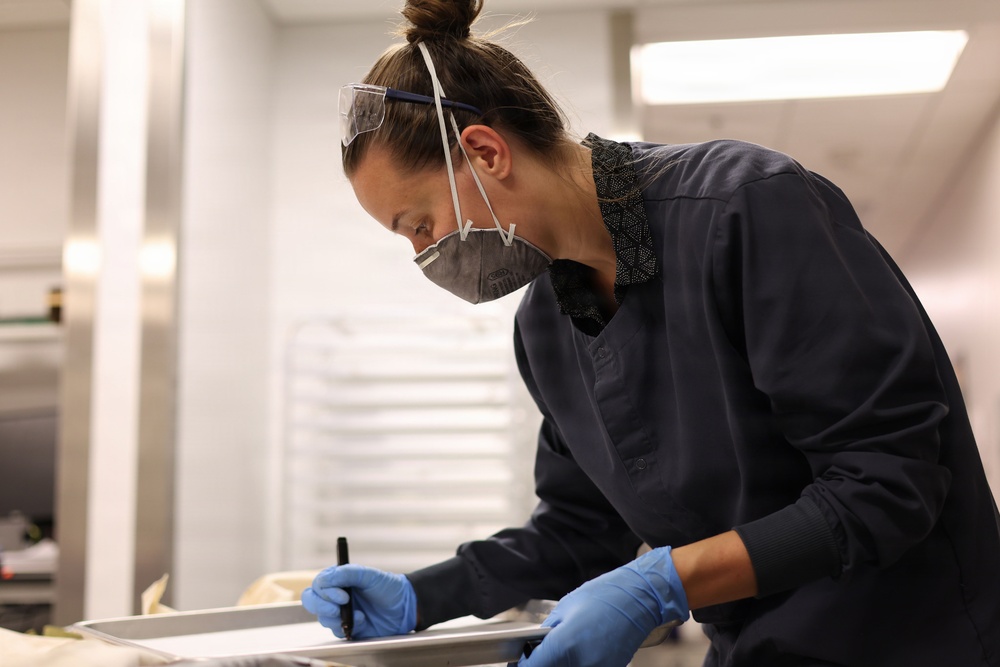 A professional, likely a scientist, medical examiner, or laboratory technician, is leaning over a work surface and writing on a document or form. She is wearing full personal protective equipment including a respirator mask, safety goggles pushed up on her forehead, blue nitrile gloves, and dark navy scrubs. The setting appears to be a clinical or forensic laboratory environment.
