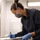 A professional, likely a scientist, medical examiner, or laboratory technician, is leaning over a work surface and writing on a document or form. She is wearing full personal protective equipment including a respirator mask, safety goggles pushed up on her forehead, blue nitrile gloves, and dark navy scrubs. The setting appears to be a clinical or forensic laboratory environment.