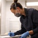 A professional, likely a scientist, medical examiner, or laboratory technician, is leaning over a work surface and writing on a document or form. She is wearing full personal protective equipment including a respirator mask, safety goggles pushed up on her forehead, blue nitrile gloves, and dark navy scrubs. The setting appears to be a clinical or forensic laboratory environment.