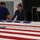 Two individuals are working with a large American flag draped over a flat surface in what appears to be an indoor facility. One person is wearing a protective face mask while both appear to be carefully handling or folding the flag. The setting suggests a professional or institutional environment, possibly related to flag preparation or ceremonial duties.
