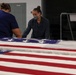 Two individuals are working with a large American flag draped over a flat surface in what appears to be an indoor facility. One person is wearing a protective face mask while both appear to be carefully handling or folding the flag. The setting suggests a professional or institutional environment, possibly related to flag preparation or ceremonial duties.