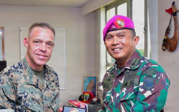 Two military personnel are posing together while jointly holding a commemorative plaque or medallion award. One individual wears a U.S. Marine Corps MARPAT digital camouflage uniform with a name tape reading 'DETRINIA,' while the other wears an Indonesian Armed Forces (TNI AL - Tentara Nasional Indonesia Angkatan Laut) camouflage uniform with a distinctive magenta/pink beret. The setting appears to be a military office, suggesting a formal bilateral exchange or ceremony.