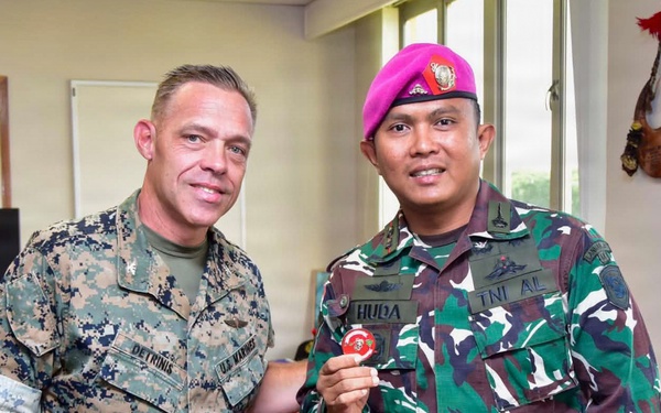 Two military personnel are posing together in an office setting, with one holding a red challenge coin or medallion toward the camera. The individual on the left wears U.S. Marines digital camouflage (MARPAT) with a name tape reading 'DETRINIS' and lieutenant colonel oak leaf insignia. The individual on the right wears Indonesian military (TNI AL - Tentara Nasional Indonesia Angkatan Laut/Navy) camouflage uniform with a pink/magenta beret, name tape reading 'HUDA', and various unit patches.