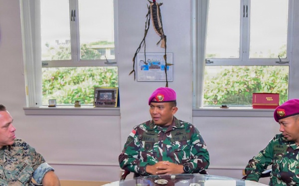 Three military personnel are seated around a glass-topped round table in what appears to be an office or meeting room. One individual wears U.S. Marine Corps digital camouflage (MARPAT), while the other two wear Indonesian Armed Forces (TNI) woodland camouflage uniforms with distinctive magenta/maroon berets, indicative of the Korps Marinir (Indonesian Marine Corps). They appear to be engaged in a formal bilateral military discussion.